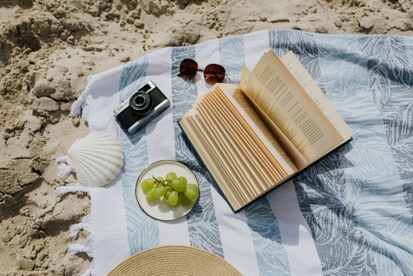 Flat lay of book, camera, and grapes on a beach towel by the sand.